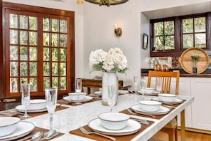 A dining room with a long table set for six, a vase of white flowers, and a view of the outdoors through large windows.