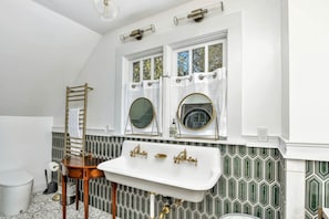Un baño con un lavamanos blanco, dos espejos, un perchero para toallas y una ventana con cortinas.