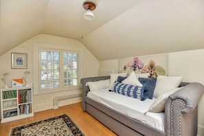 A bedroom with a grey sofa bed, a bookshelf, a window with blinds, and a ceiling light fixture.