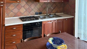 A kitchen with wooden cabinets, a tiled backsplash, a stove, a sink, and a dining table with a blue and yellow bowl. - Oasis aloe vera (Troina)