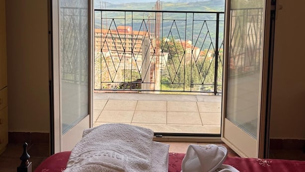 A balcony with a view of a cityscape, a bed with a red bedspread, and a pair of white slippers. - Oasis aloe vera (Troina)