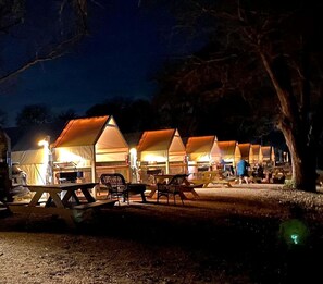 A row of tents with tables and chairs, set up in a campsite at night.