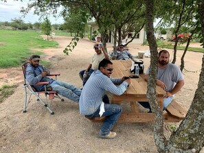 A group of people seated around a wooden table outdoors, with trees and a grassy field in the background.