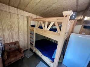 A wooden bunk bed with a blue mattress, a small wooden chair, and a white refrigerator in a room with wooden walls and a sloped ceiling.