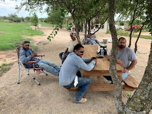 A group of people seated around a wooden table outdoors, with trees and a grassy field in the background.