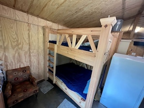 A wooden bunk bed with a blue mattress, a small wooden chair, and a white refrigerator in a room with wooden walls and a sloped ceiling.