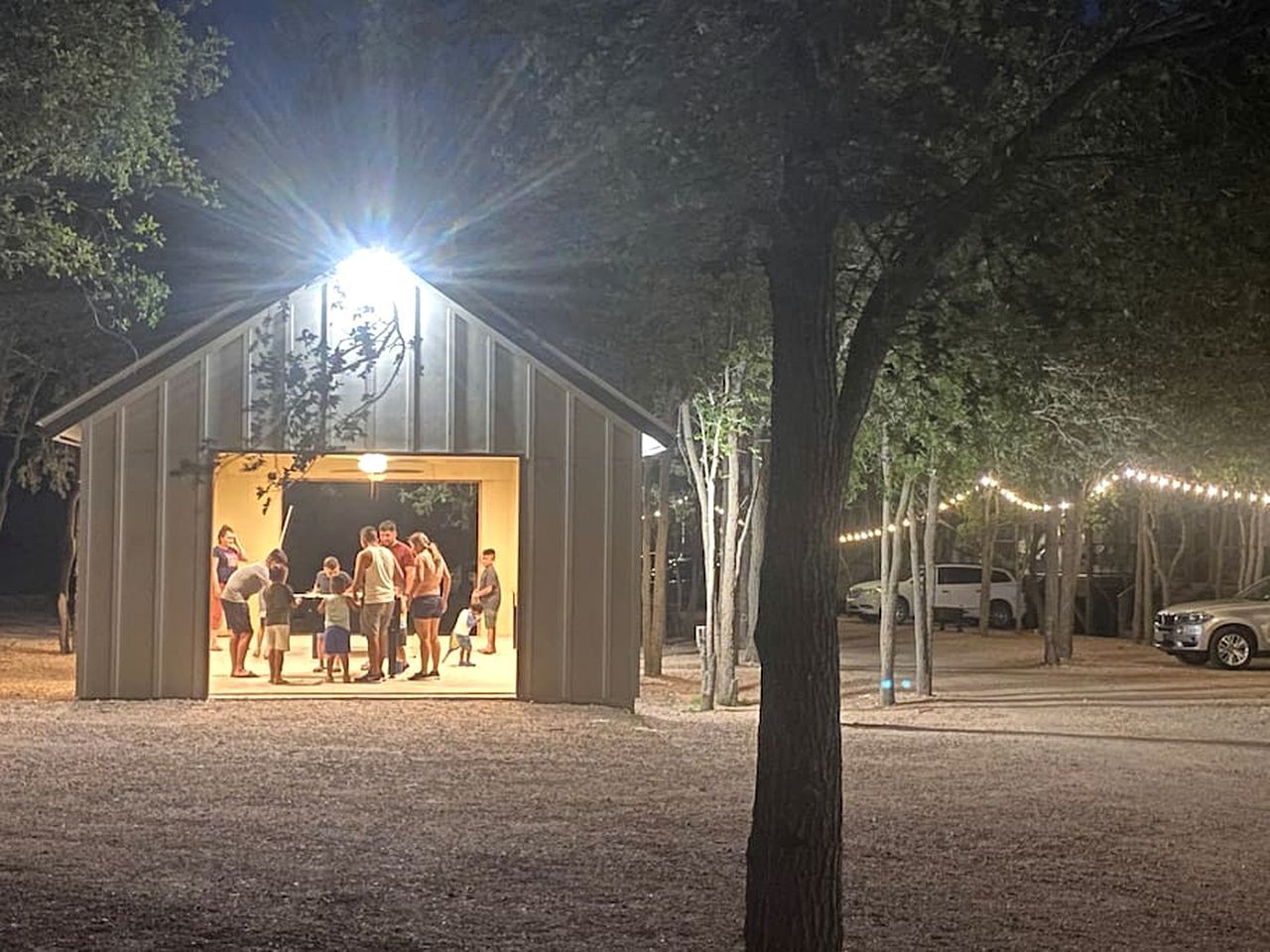 Een overdekt buitengebied waar een groep mensen bij elkaar is, omringd door bomen en lichtslingers.