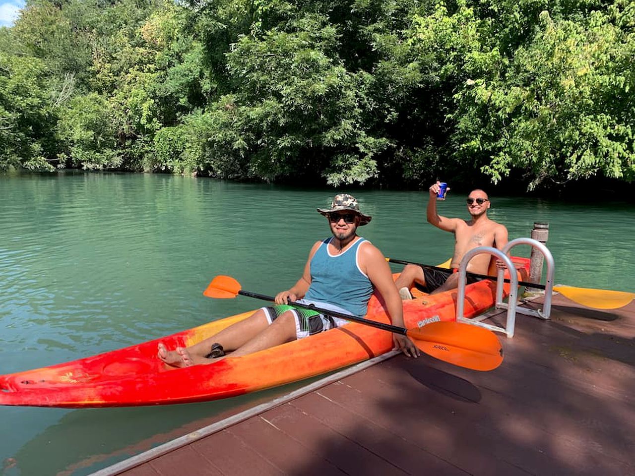 Zwei Personen in einem Kajak an einem Steg, im Hintergrund ein von Bäumen gesäumter Fluss.