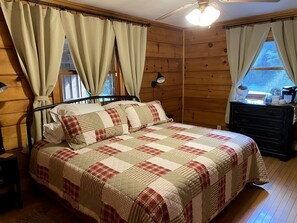 Habitación con paneles de madera, una cama, un aparador, una lámpara y una ventana con cortinas.