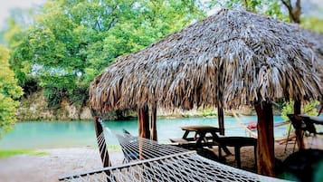 Una estructura con techo de paja, con una hamaca y una mesa de picnic junto a un cuerpo de agua tranquilo, rodeada de follaje.