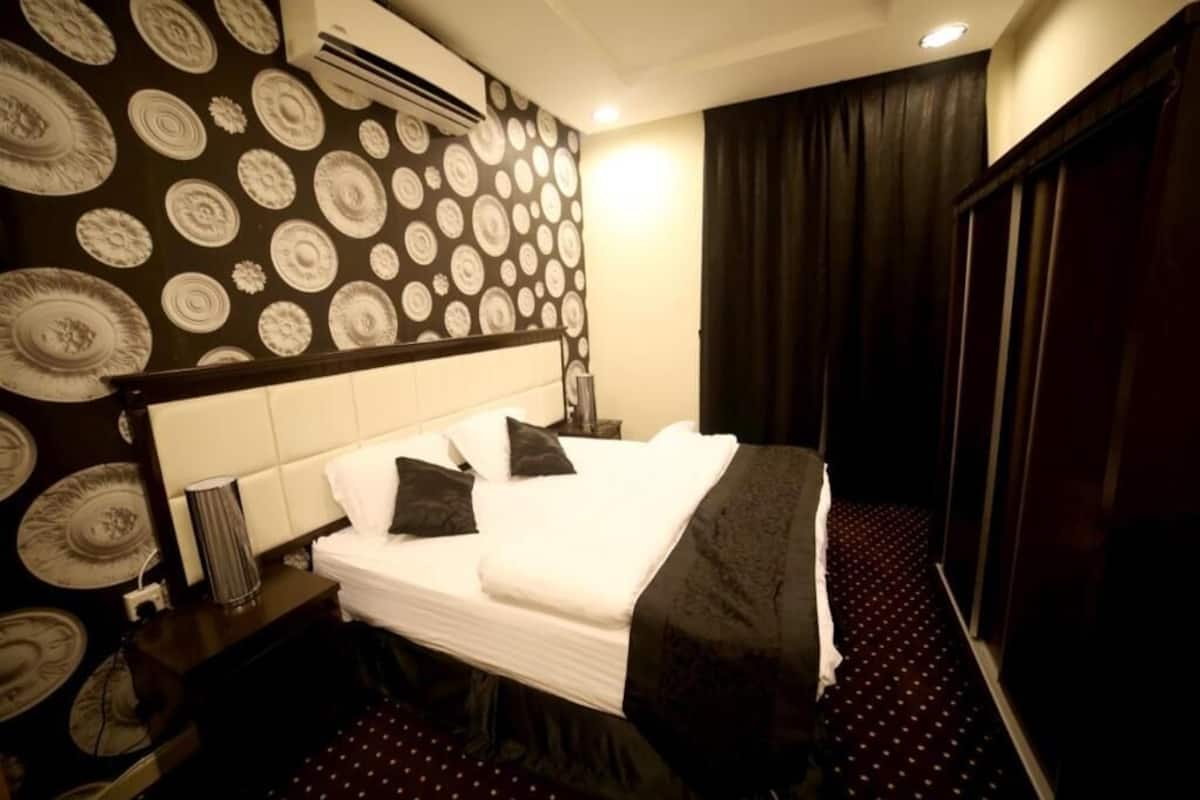 A hotel room with a large bed, a headboard, a bedside table, a lamp, a wall with circular patterns, and a dark curtain.