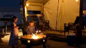 Een tent met een vuurkuil ervoor, omringd door stoelen en tafels.
