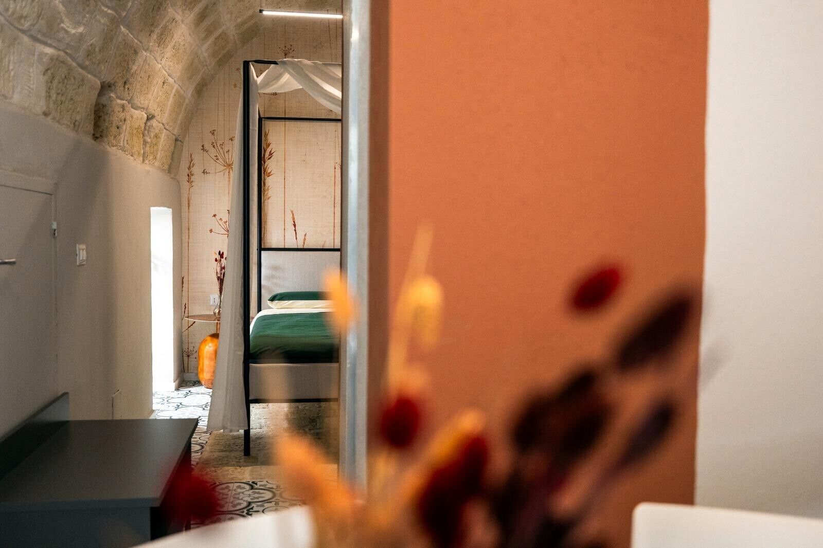 A hotel room with a stone wall, a bed with green bedding, a small table, and a vase with dried flowers.