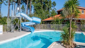 A swimming pool with a blue slide, surrounded by a fenced area, trees, and a building with a red roof. - Nature Stay Near Biscarrosse Lake (Parentis-en-Born)