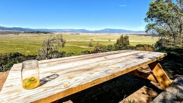 Una mesa de picnic de madera con un tarro de limonada, frente a un campo de césped y montañas distantes.