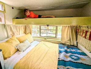 Una cama litera con un niño en la cama superior, una ventana con cortinas y una cama con ropa de cama amarilla.