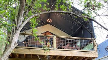 A wooden balcony with a metal railing, a black awning, and a tree in the foreground.