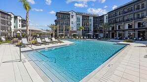 Una piscina con sillas de descanso y sombrillas, rodeada de edificios de varios pisos con balcones.