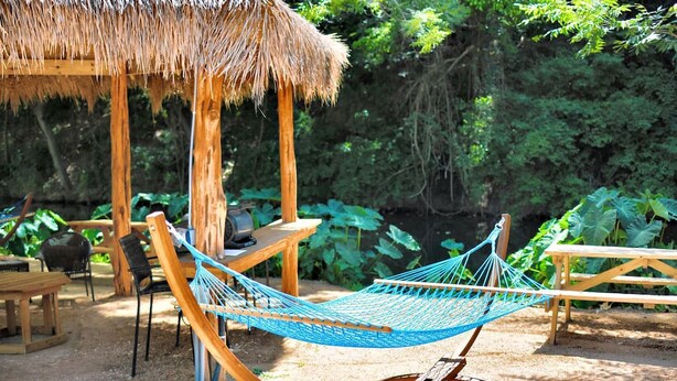 A hammock, wooden chairs, a table, and a thatched-roof structure in a garden setting.