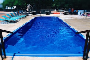 Una piscina rectangular con un tono azul, rodeada de sillas de descanso y una zona con sombra.
