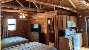 A cabin-style room with a wooden ceiling, a bed, a TV, a kitchenette, and a door leading to another room. - A Rustic Log Cabin - Perfect for a Glamping Getaway by the San Marcos River near Kingsbury, Texas (Kingsbury)