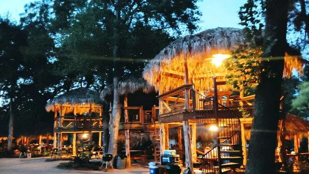A two-story wooden structure with a thatched roof, surrounded by trees, and equipped with outdoor seating and a barbecue area. - A Rustic Log Cabin - Perfect for a Glamping Getaway by the San Marcos River near Kingsbury, Texas (Kingsbury)