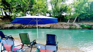 A beach with chairs and a blue umbrella. - A Rustic Log Cabin - Perfect for a Glamping Getaway by the San Marcos River near Kingsbury, Texas (Kingsbury)