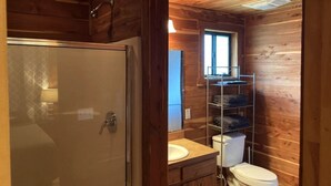 A wooden cabin bathroom with a glass shower door, a toilet, a sink, and a shelf with folded towels. - A Rustic Log Cabin - Perfect for a Glamping Getaway by the San Marcos River near Kingsbury, Texas (Kingsbury)