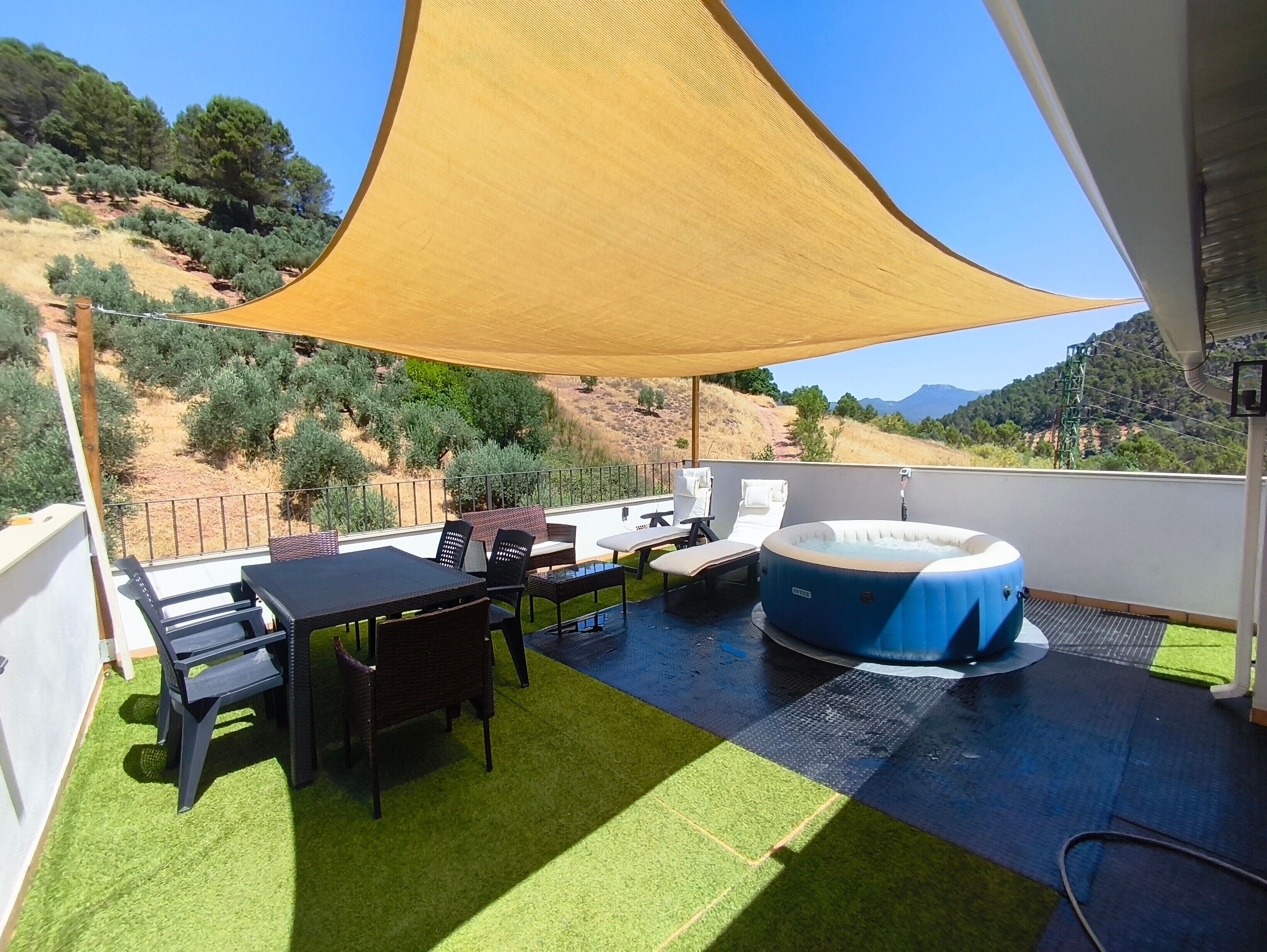 A rooftop terrace with a table, chairs, a hot tub, and a view of a hilly landscape.