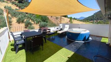 A rooftop terrace with a table, chairs, a hot tub, and a view of a hilly landscape.