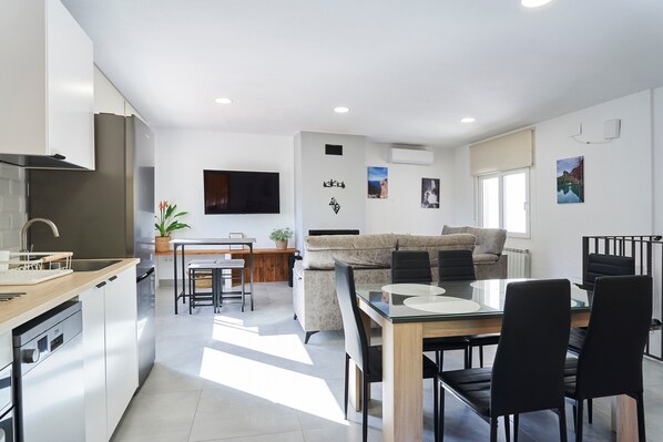 A modern kitchen with a dishwasher, a dining area with a glass table and chairs, a sofa, and a TV mounted on the wall.