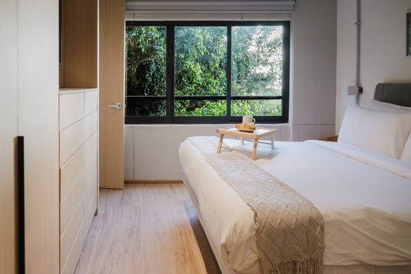 A bedroom with a bed, a window with a view of greenery, a small table with a cup and a pastry, and a wardrobe. - Modern 1BR in Juárez with fast WiFi, great natural light, a comfortable king bed, a fully equipped kitchen, and a luxury bathroom. Perfect for couples or solo travelers seeking comfort, style, and a prime location in Mexico City. (Cuauhtémoc)