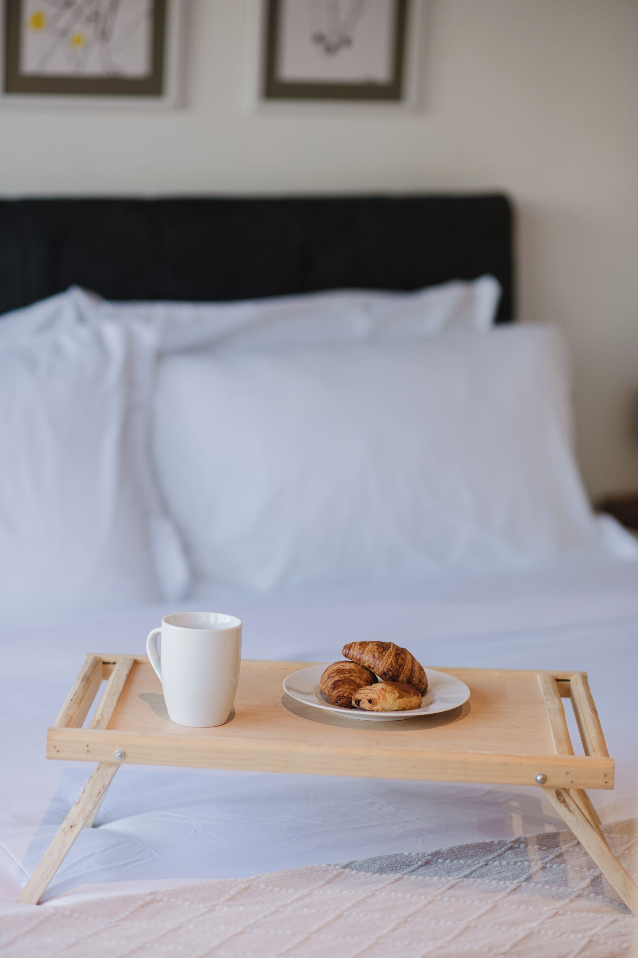 Una cama con ropa blanca, una bandeja de madera con una taza blanca y croissants, y un cuadro en la pared.