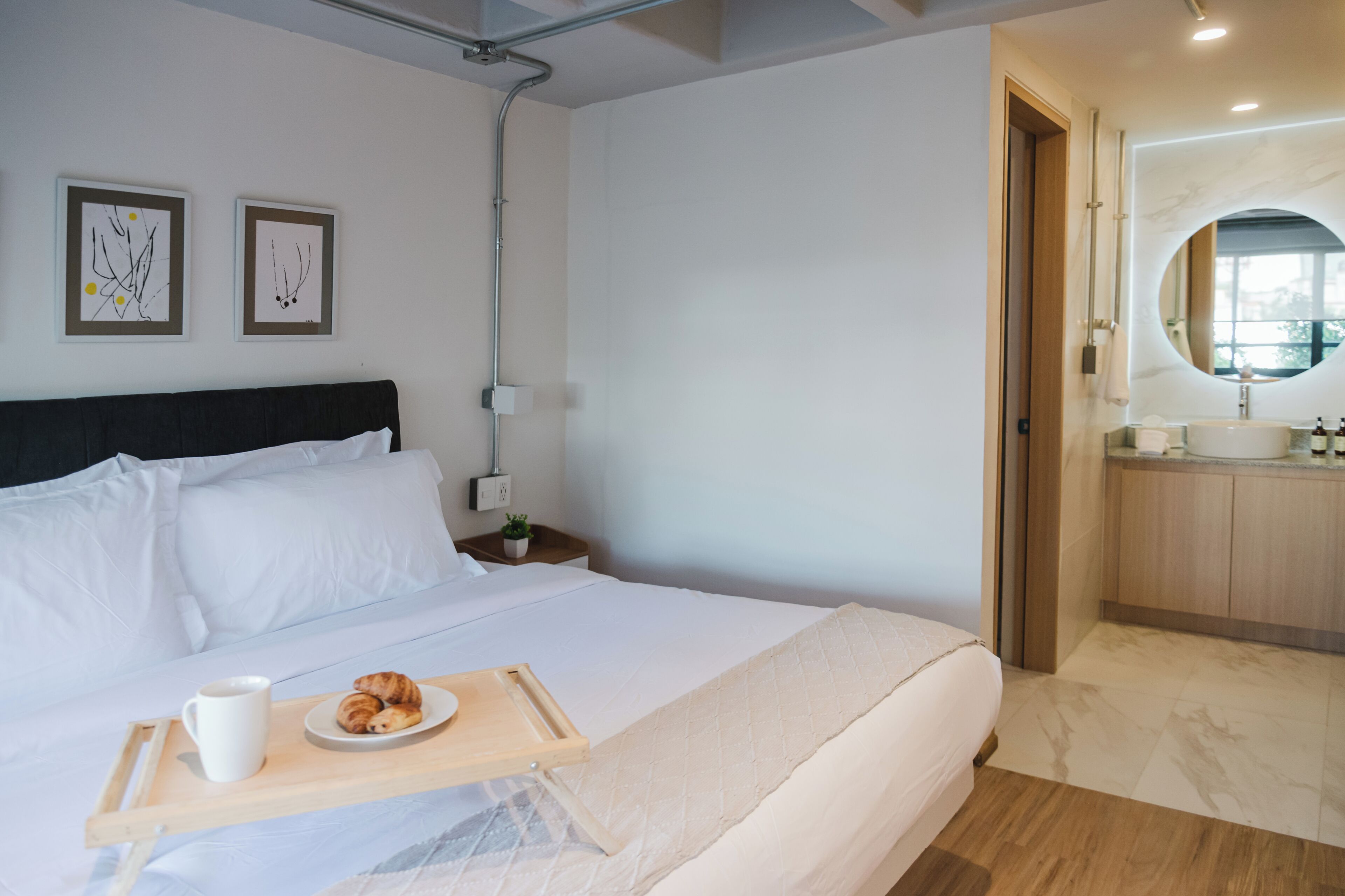 Une chambre d’hôtel avec un lit, un plateau avec des croissants et une tasse, une table de chevet avec une plante, et une salle de bain avec un miroir et un lavabo.