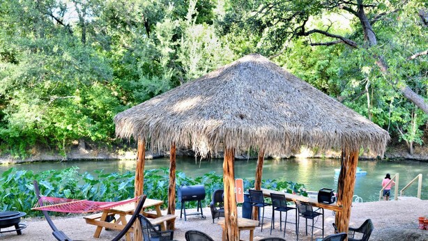 A thatched-roof pavilion with outdoor seating and a grill, surrounded by greenery and a body of water. - Cabaña Son's Rio Cibolo Waterfront Cabin, San Antonio, Texas (Marion)