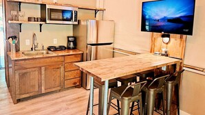 A compact kitchenette with a microwave, refrigerator, and coffee maker. A wooden table with metal chairs is positioned in front of the counter. A flat-screen TV is mounted on the wall above the counter. - Cabaña Son's Rio Cibolo Waterfront Cabin, San Antonio, Texas (Marion)