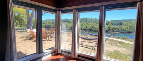 A room with large windows, wooden ceiling, and a view of a lakeside area with outdoor furniture.