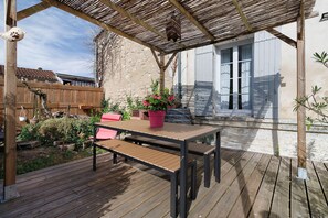 Um deck de madeira com mesa e bancos, cercado por um jardim e um edifício com porta branca.