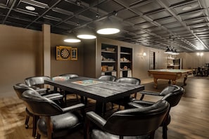 Quarto com mesa de bilhar, poltronas de couro, alvo de dardos, prateleiras com cestos e teto com vigas aparentes.
