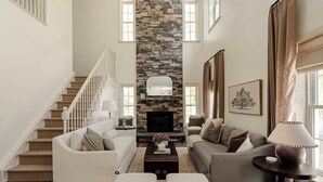 A spacious living room with a stone fireplace, a staircase, and a large window.