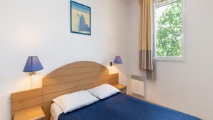 A bedroom with a wooden headboard, two bedside tables with lamps, a bed with a blue bedspread, a window with curtains, and a framed picture on the wall. - 6-guest Flat in Landes Forest (Ondres)