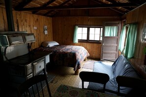 Interior rústico de una cabaña con una cama, un escritorio con estufa, una silla y una ventana con cortinas.