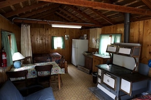 A rustic cabin with a wooden interior, a dining table with chairs, a kitchenette with a stove, and a refrigerator.