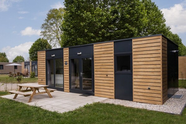 A modern, single-story building with a wooden exterior and large glass doors, situated in a grassy area with a picnic table in front.