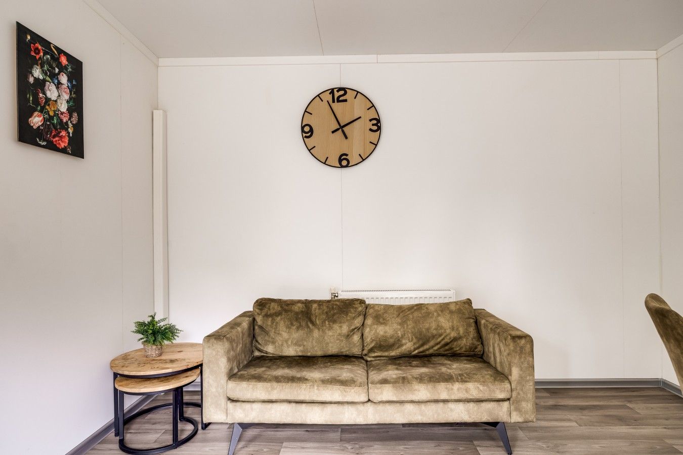 A living room with a beige sofa, a round wooden clock, a small round table with a plant, and a radiator on the wall.