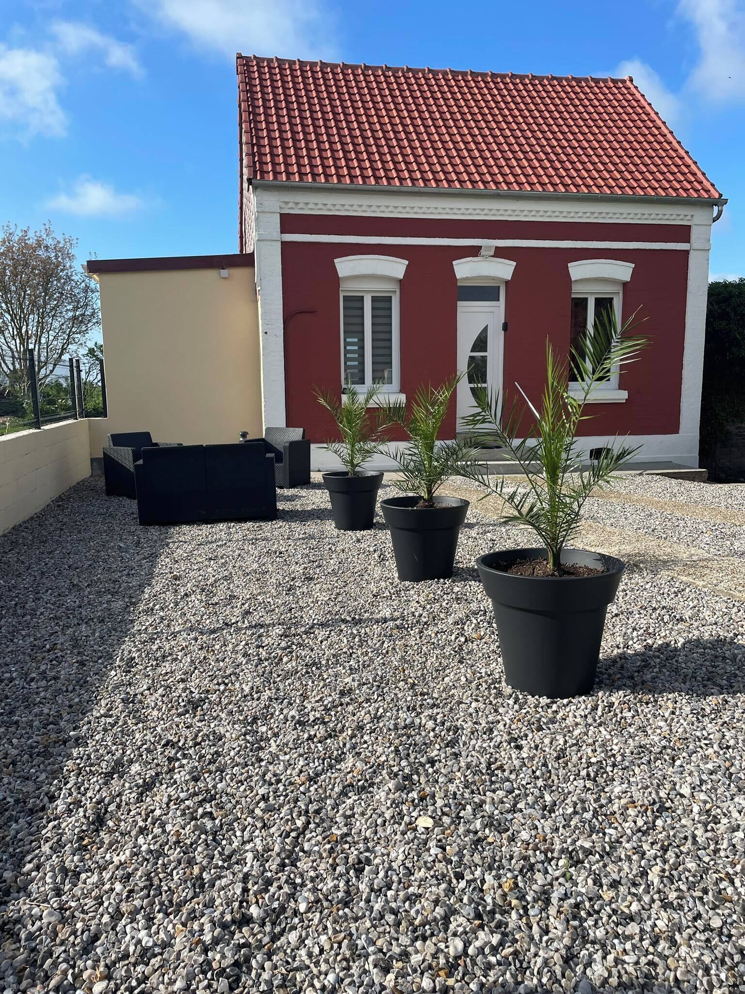 Un bâtiment avec un toit rouge et des éléments décoratifs blancs, une terrasse en gravier et des plantes en pot.