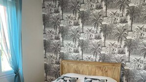 Une chambre à coucher avec un lit, une fenêtre avec des rideaux, un mur recouvert d’un papier peint à motifs tropicaux et un luminaire suspendu.