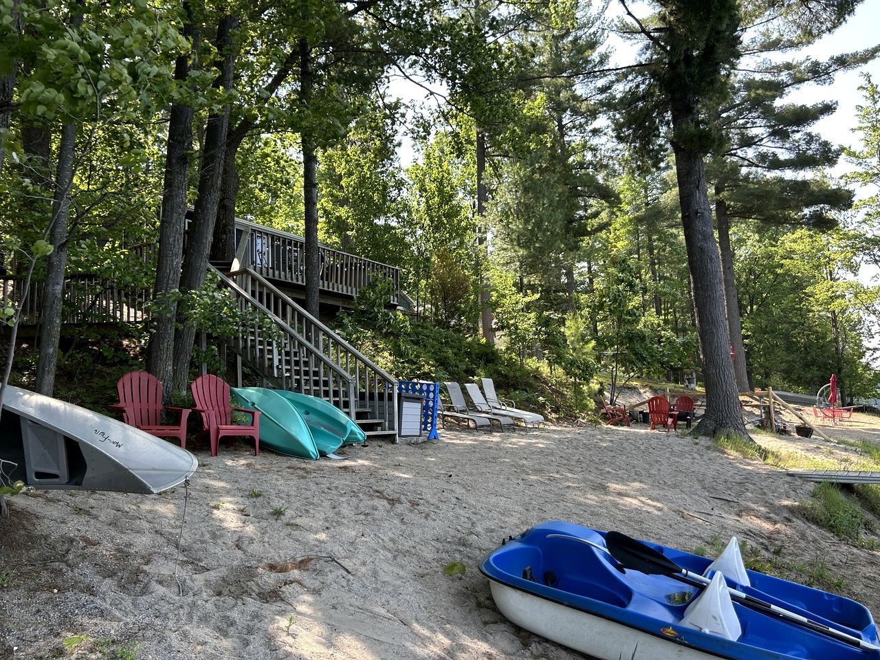 A beach area with a staircase leading to a deck, surrounded by trees. There are beach chairs, a kayak, and a surfboard.