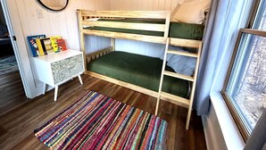 Quarto com beliche, uma mesa consola branca, tapete listrado e uma janela com cortinas.