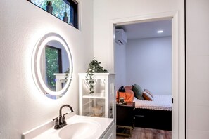 Une salle de bain avec un miroir rond, un lavabo, une plante en pot et un lit dans la pièce adjacente.